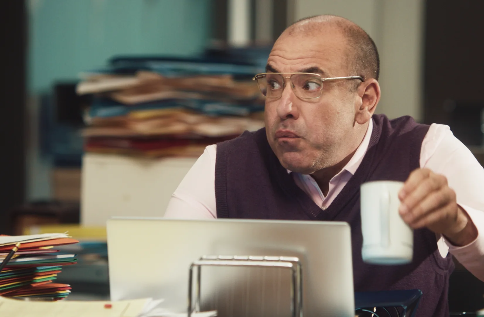 A man wearing glasses and a purple sweater vest sits at a cluttered desk, holding a white mug and looking to his right with a surprised expression. A laptop and piles of colorful folders are in front of him.