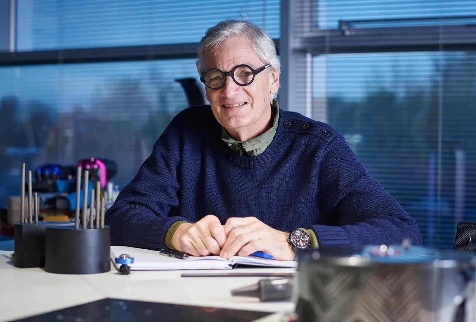 An older man with gray hair and round glasses, wearing a navy sweater, sits at a desk with papers and office supplies in front of him, smiling in a modern office with large windows.