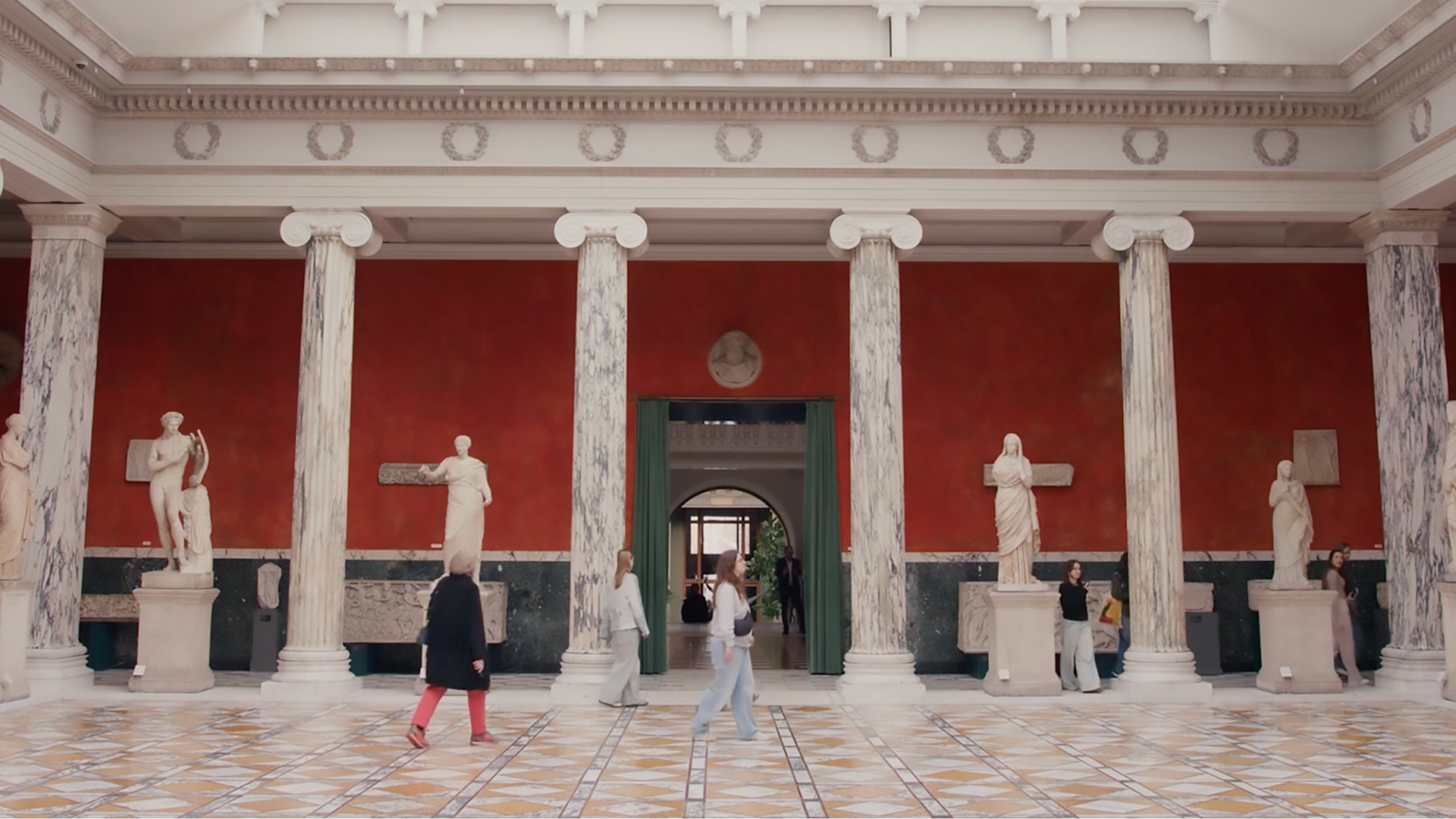 A museum hall with red walls, white columns, and statues. People walk on a patterned floor.