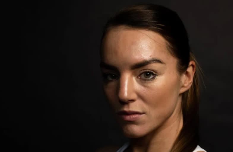 A woman with brown hair pulled back poses against a dark background, looking seriously into the camera. She is lit from the front, highlighting her facial features.