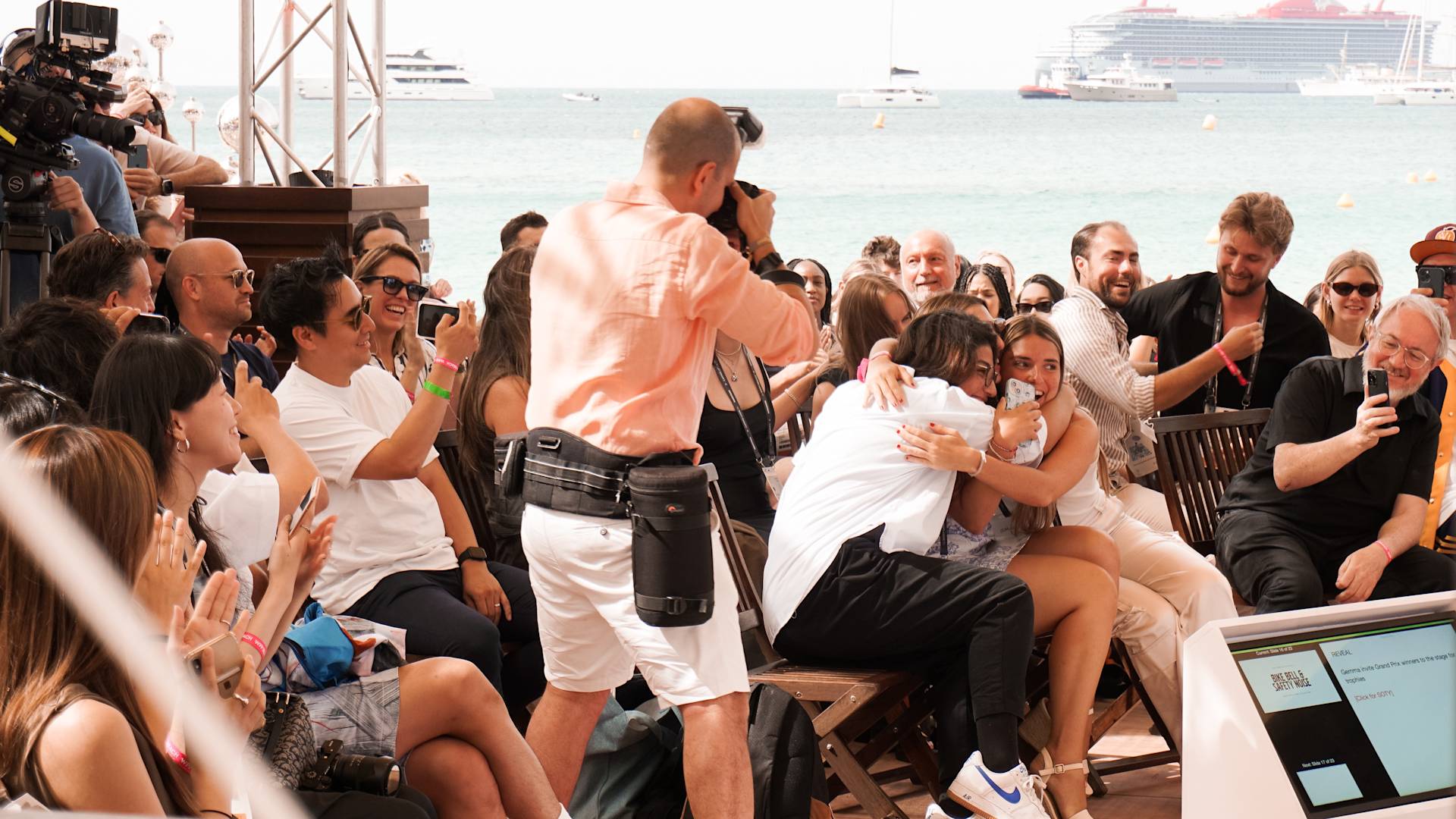 A crowd sits and stands by the seaside, applauding as two people embrace in the center, while a photographer captures the moment. Smiling faces show excitement and joy at the lively event. Yachts float in the background.