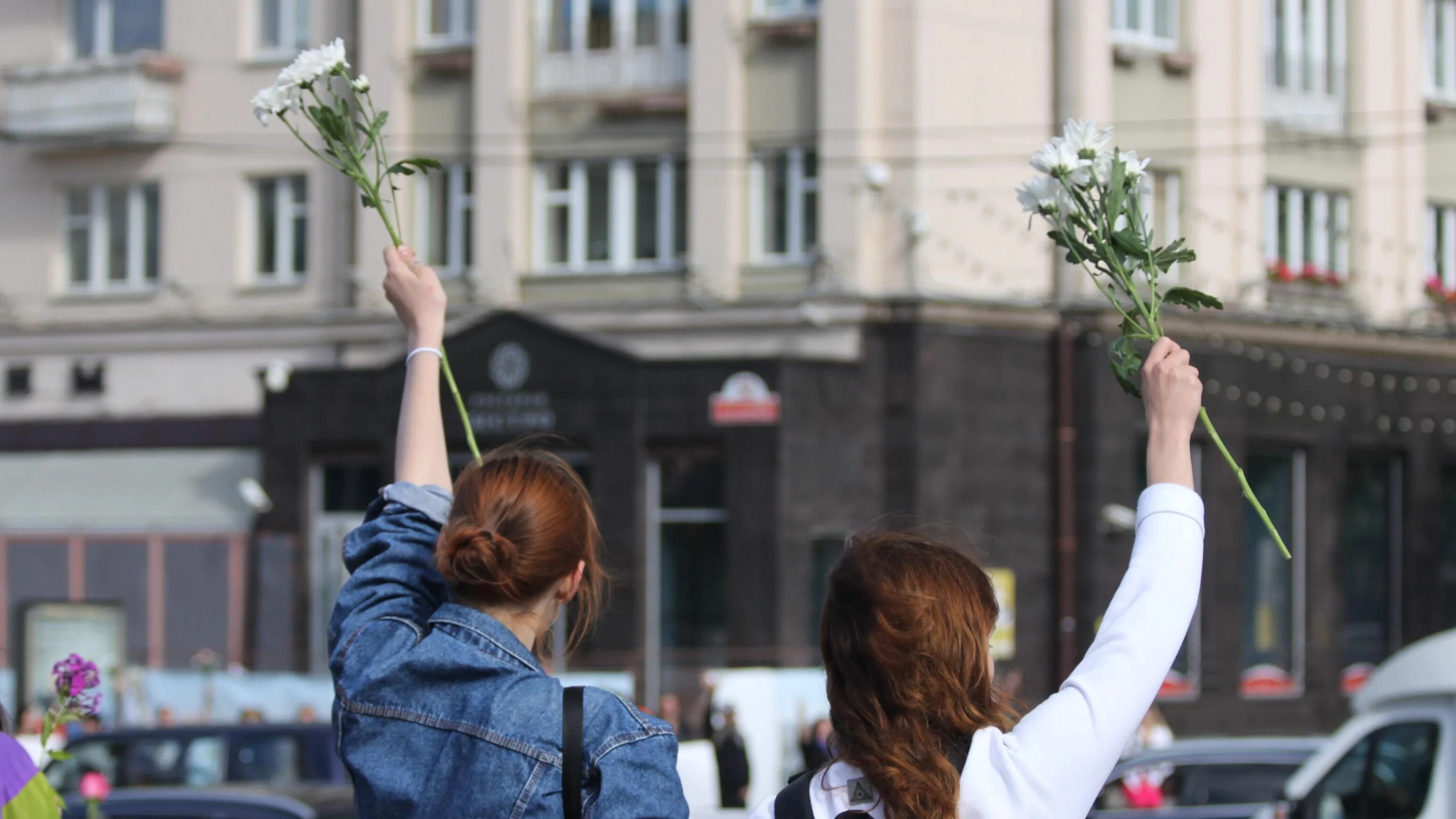 Two people with raised arms hold white flowers, facing away from the camera. They stand outdoors in front of a city building, suggesting a peaceful demonstration or gathering.