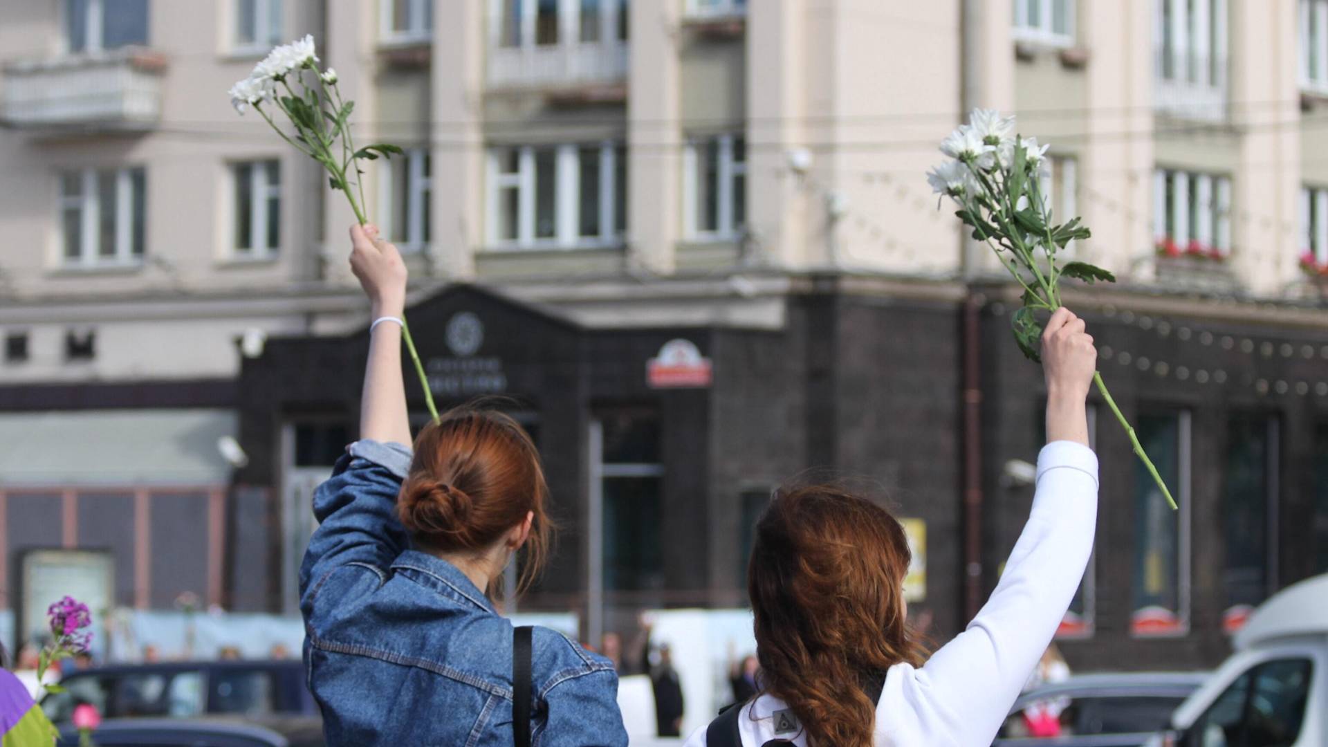 Two people with raised arms hold white flowers, facing away from the camera. They stand outdoors in front of a city building, suggesting a peaceful demonstration or gathering.