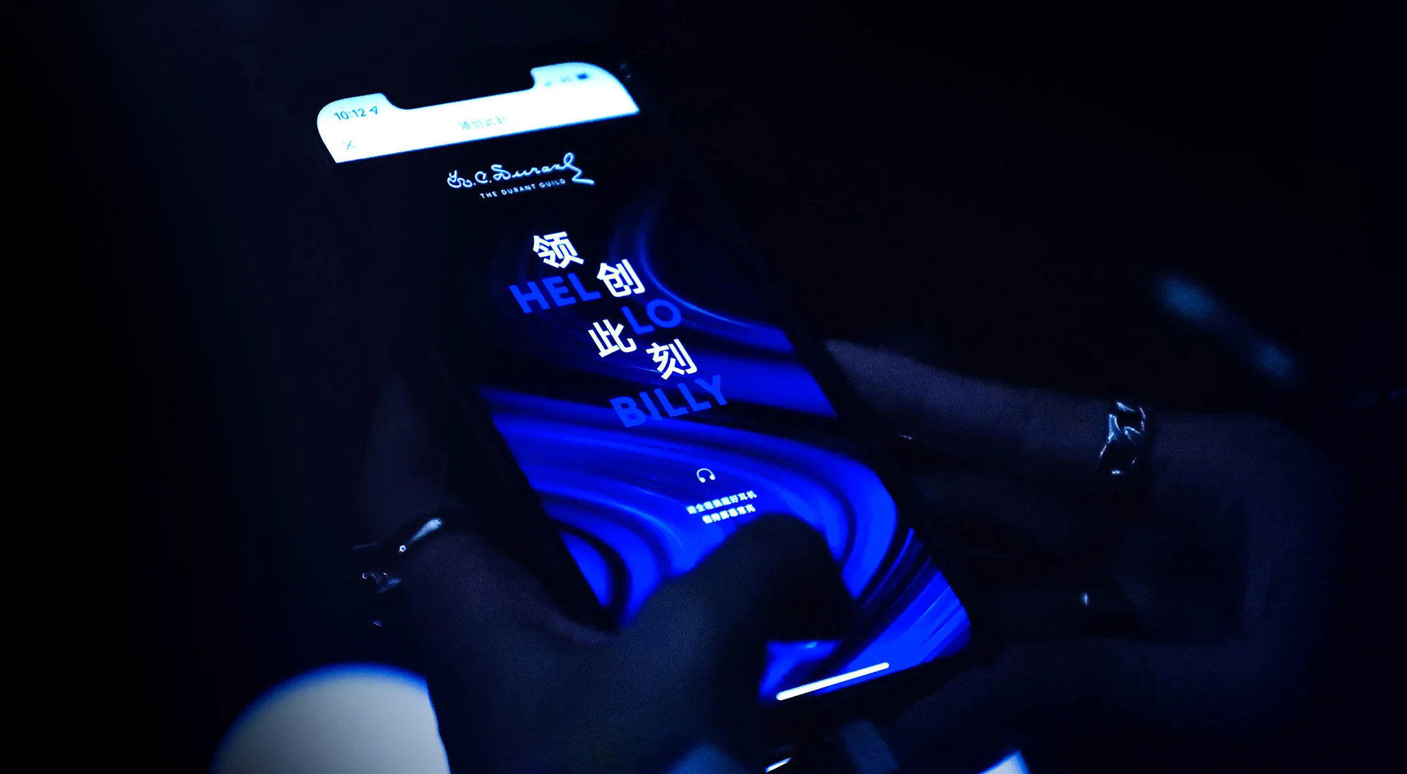 A person holding a smartphone displaying a blue and black screen with Chinese characters and the English words HELLO BILLY. The scene is dimly lit, and the person’s fingers are visible on the screen.