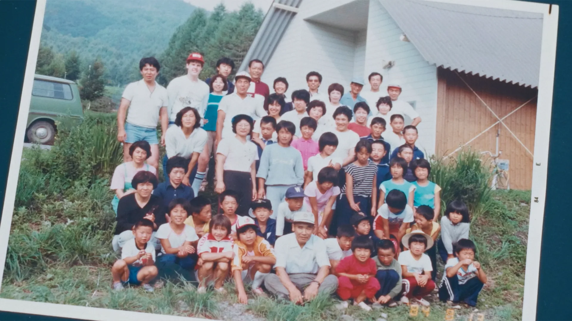 A large group of children and adults pose together outdoors in front of a light-colored building with a sloped roof, surrounded by greenery and hills, all smiling at the camera.