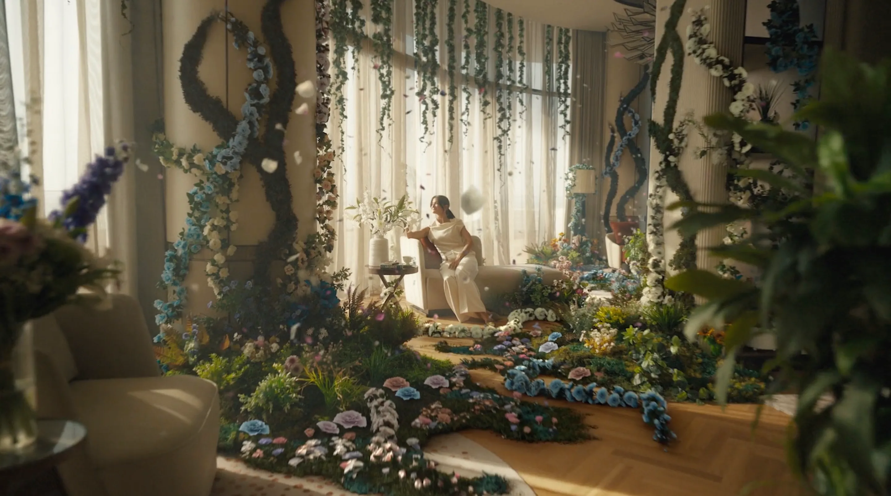 A woman in a white dress sits in a bright, elegant room filled with lush, colorful flowers and vines, which decorate the floor, columns, and ceiling, creating a whimsical, indoor garden atmosphere.