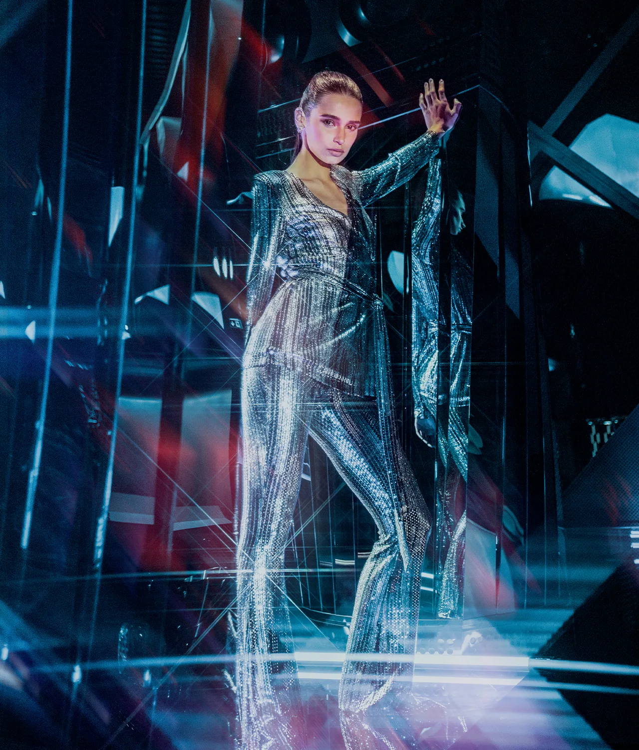 A woman in a shiny, metallic jumpsuit poses confidently with one arm raised against a futuristic, colorful background with reflective surfaces and glowing lights.