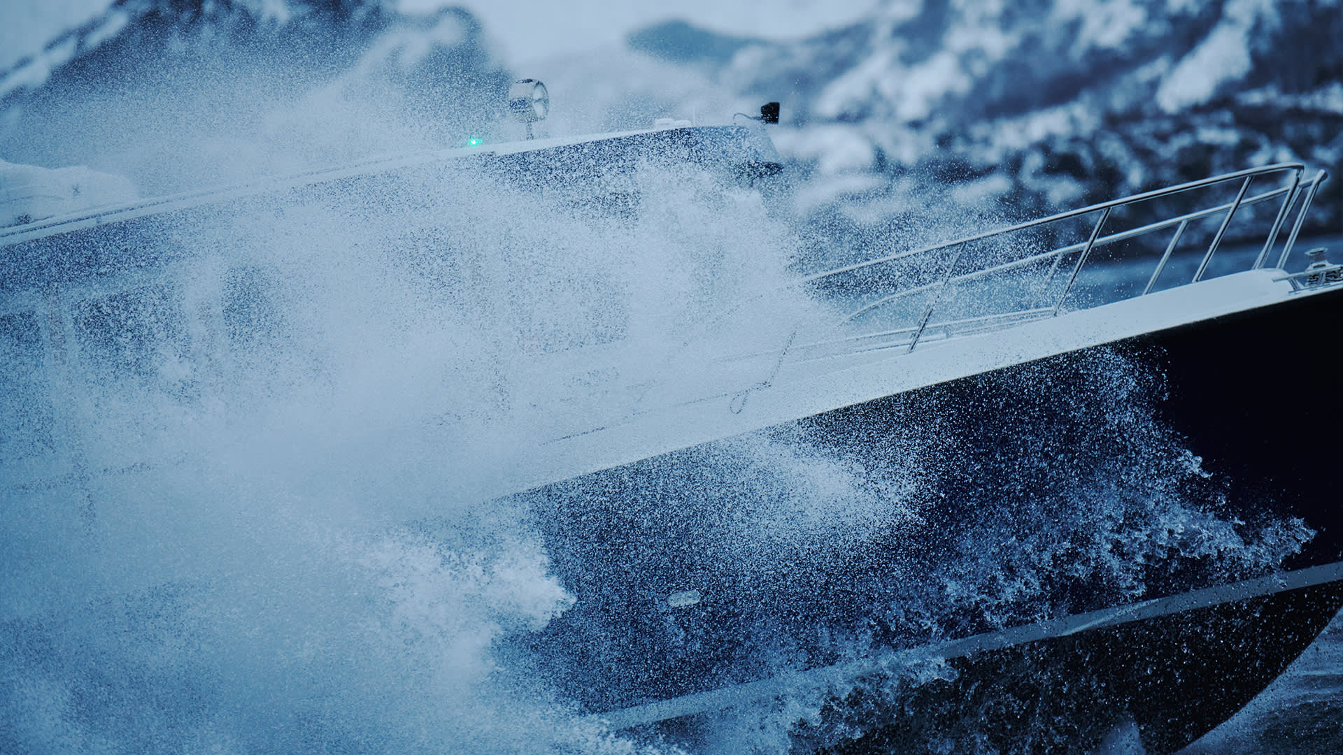A boat is cutting through rough water, creating a large spray as it moves forward, with snowy mountains blurred in the background.