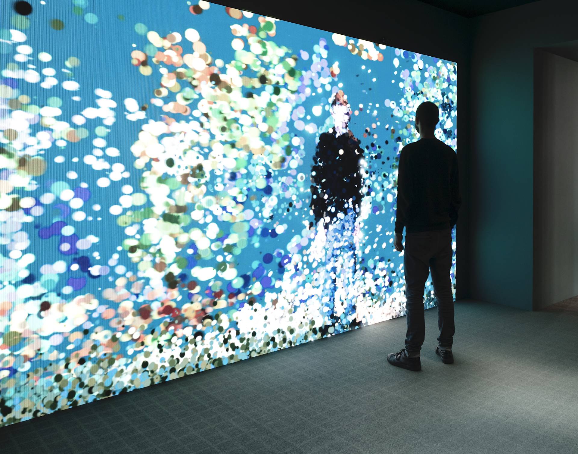 A person stands in front of a large, colorful digital screen filled with abstract, glowing dots and shapes in a dimly lit room.