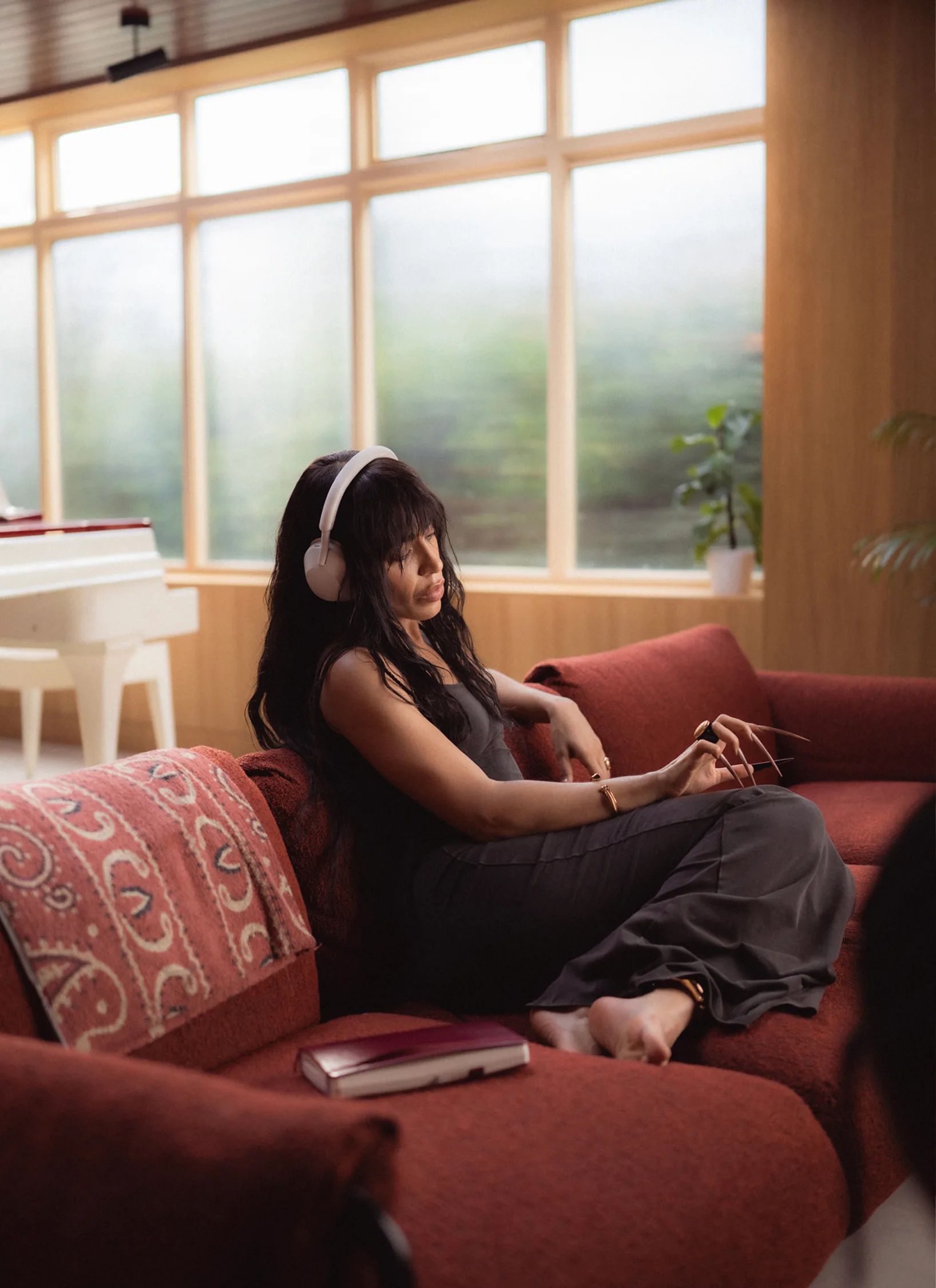 A woman with long dark hair sits barefoot on a red sofa, wearing headphones and holding a pencil. A closed book rests beside her. Large windows and a white piano are in the background.
