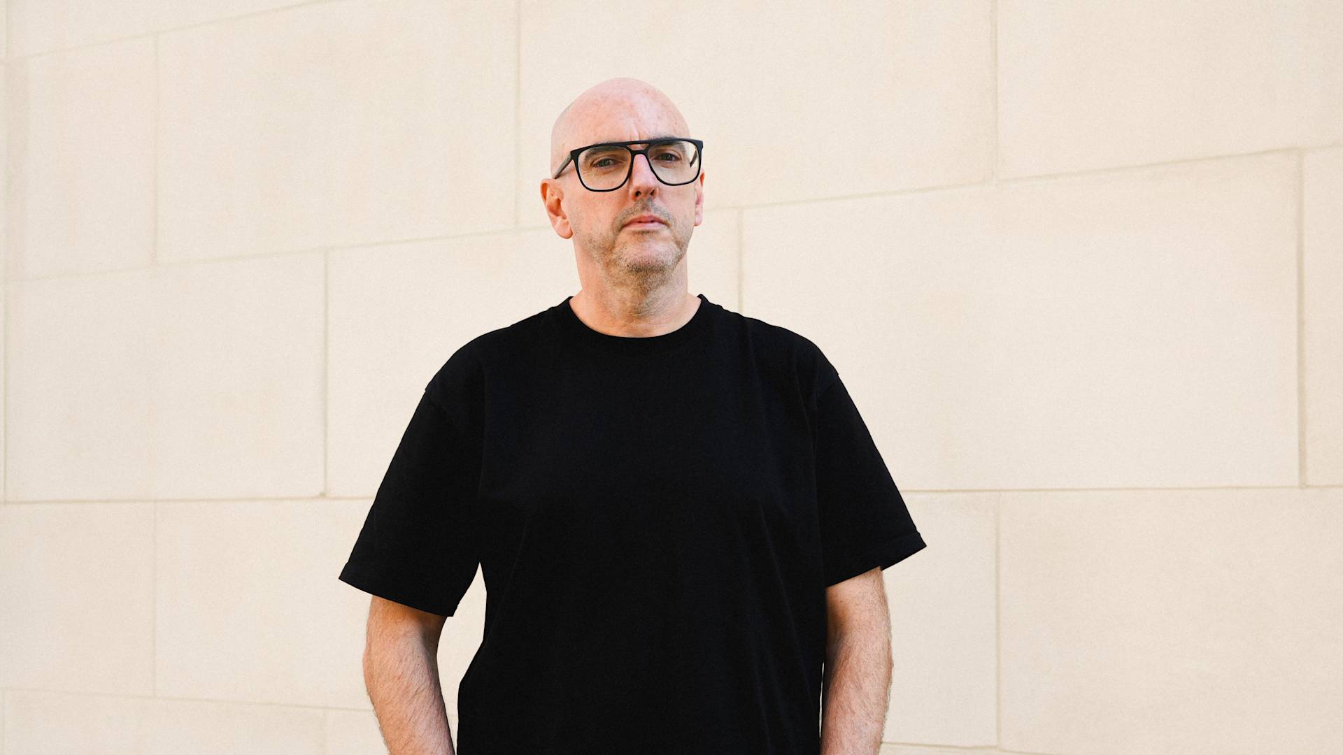 A bald man wearing black-rimmed glasses and a black t-shirt stands against a light-colored stone wall, looking directly at the camera with a neutral expression.