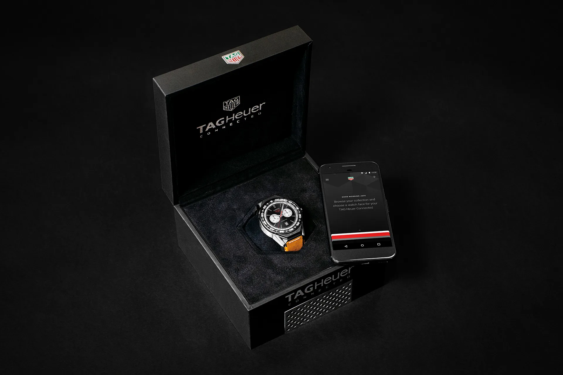 A TAG Heuer watch in an open black box is displayed next to a smartphone with a dark screen on a black background. Both the box and lid show the TAG Heuer logo.