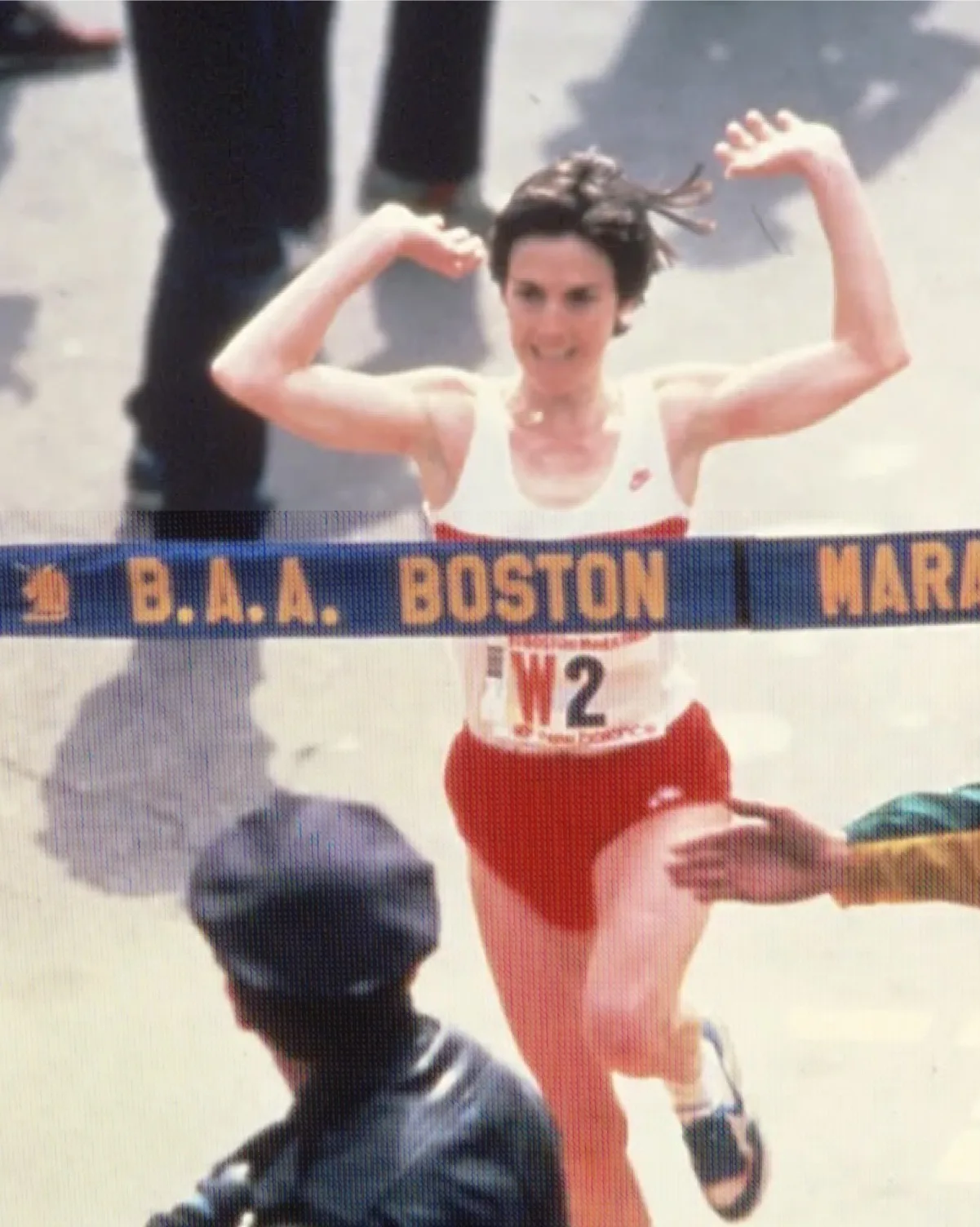 A female runner crosses the finish line of the Boston Marathon, raising her arms in celebration. She wears a white tank top and red shorts, with spectators and a police officer visible in the foreground.