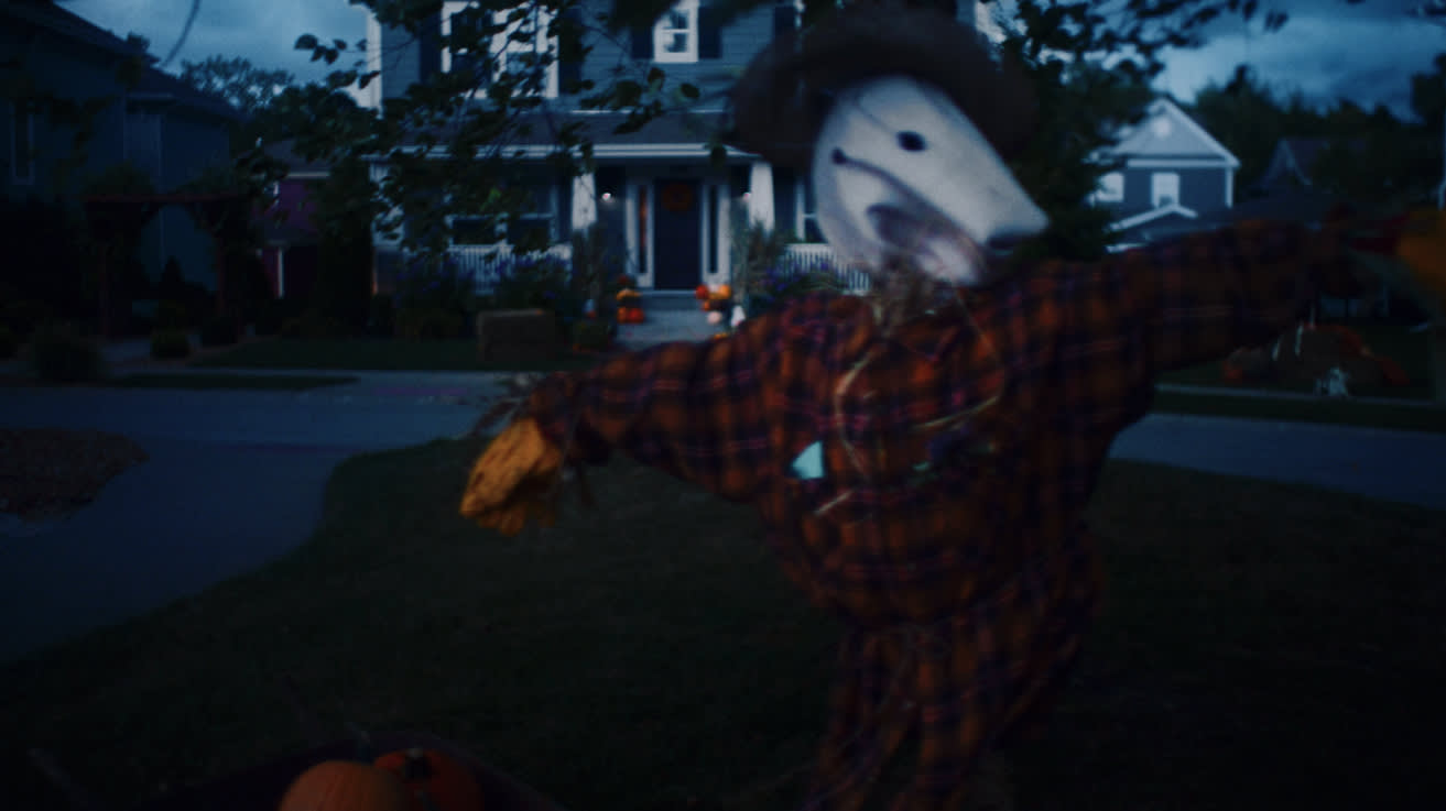 A scarecrow with a white animal mask, straw hands, and a plaid shirt stands in a yard at dusk. Pumpkins are nearby, and a house decorated for Halloween is in the background.