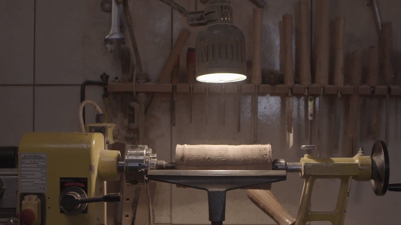 A wooden log is clamped in a lathe machine under a bright lamp in a workshop, with various woodworking tools hanging on the wall in the background.