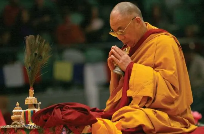 A Buddhist monk in maroon and yellow robes sits cross-legged, holding a ritual object in his hands, appearing deep in prayer or meditation. Ceremonial items are placed in front of him on a colorful cloth.