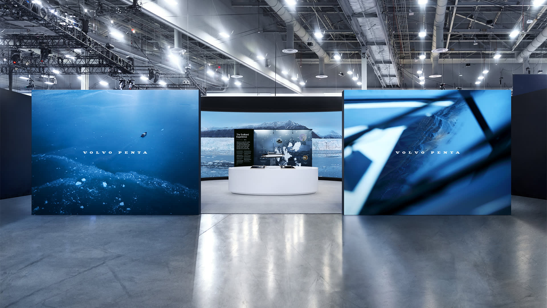 Modern exhibition booth with large blue display panels featuring the Volvo Penta logo, a central white oval kiosk, and bright overhead lighting in a spacious, industrial convention hall.