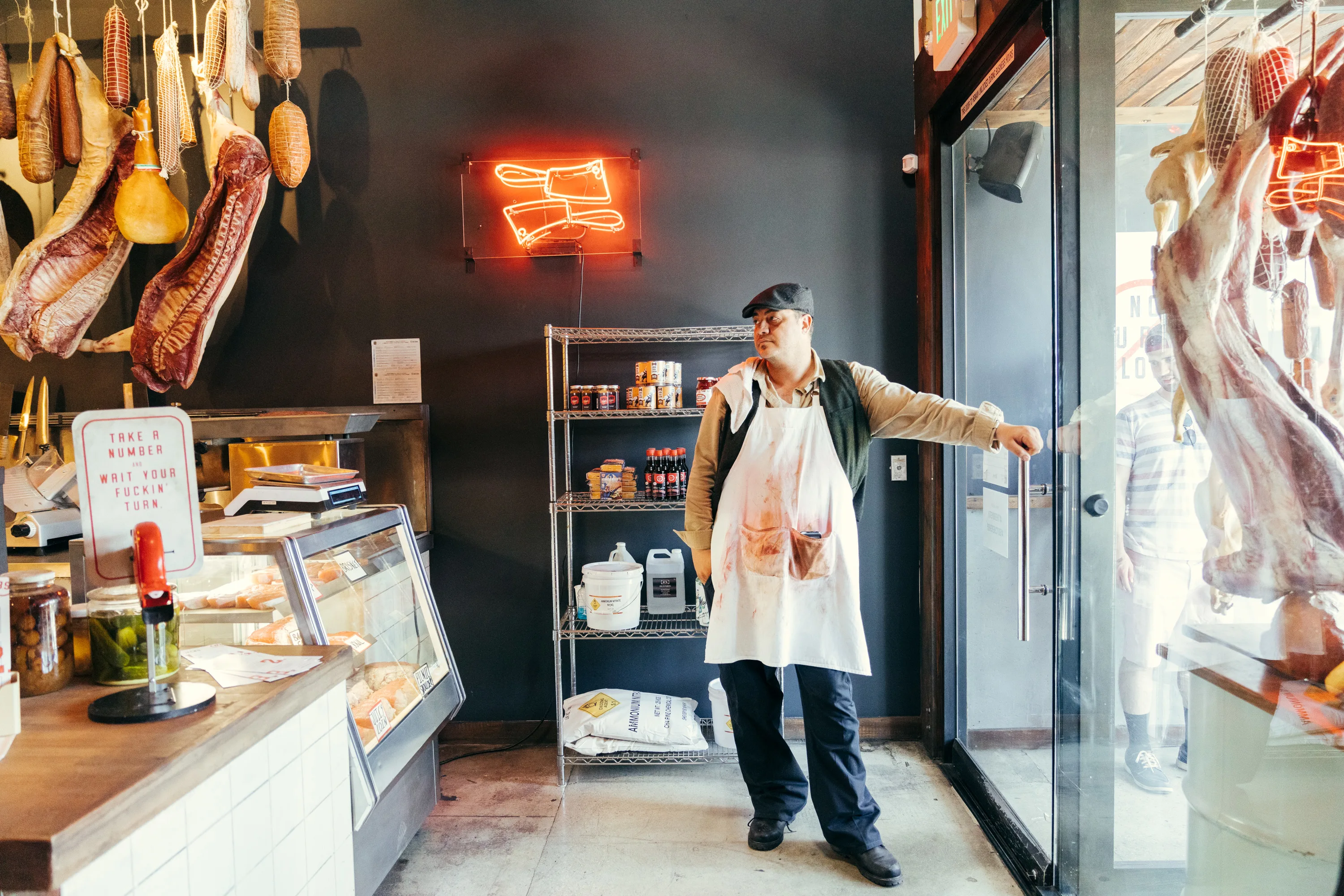 A butcher wearing an apron and cap stands by the glass door of a deli, surrounded by hanging cured meats, a display case, shelves of goods, and a red neon sign shaped like bacon.