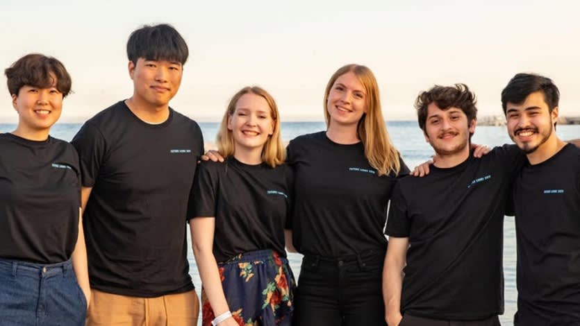 Six young adults stand in a row outdoors, smiling at the camera. They are wearing matching black t-shirts and have their arms around each other. The background shows a body of water and a light sky.