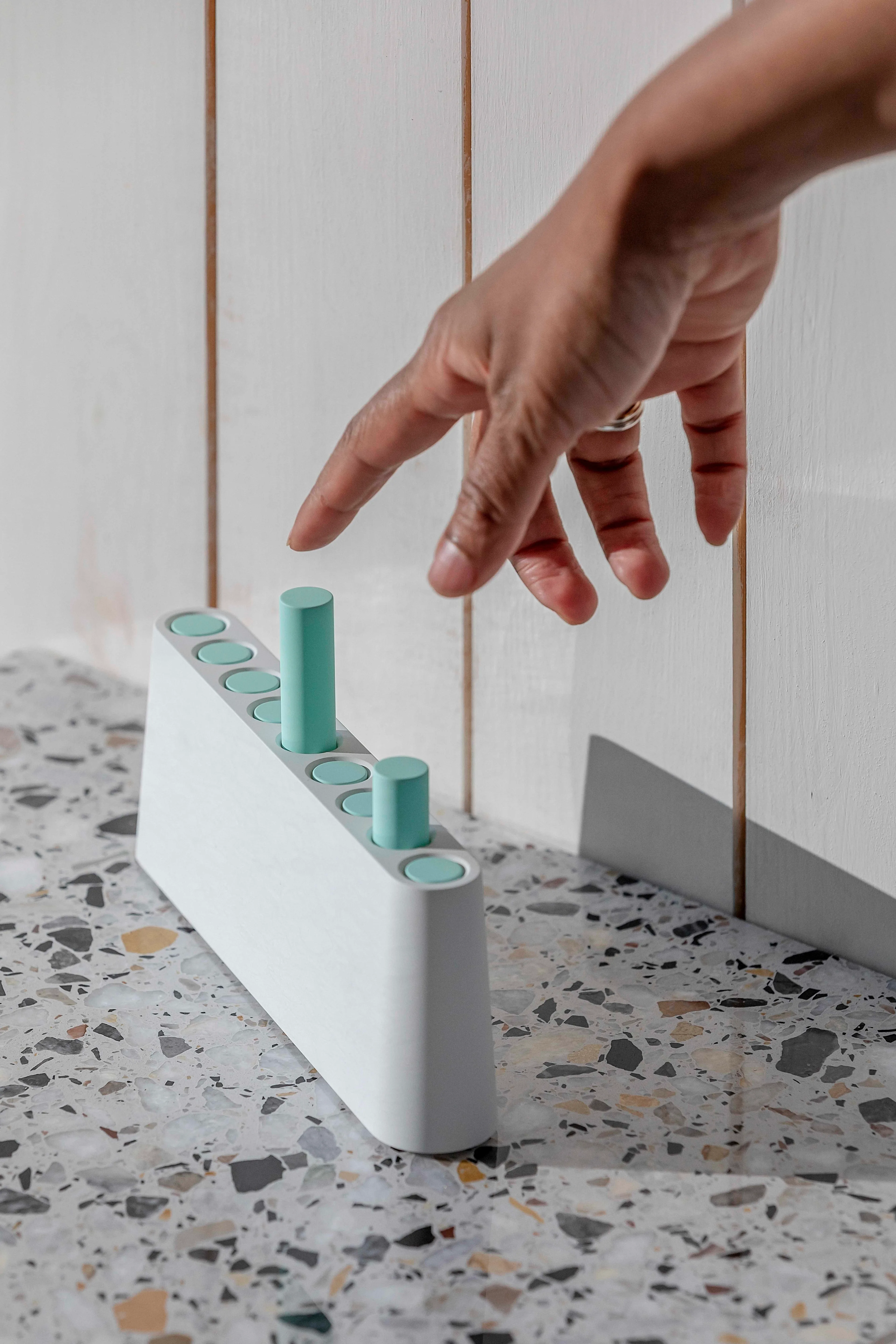 A hand reaches toward a modern, minimalist white device with several round, light blue buttons and cylindrical pegs on a speckled surface against a white wooden background.
