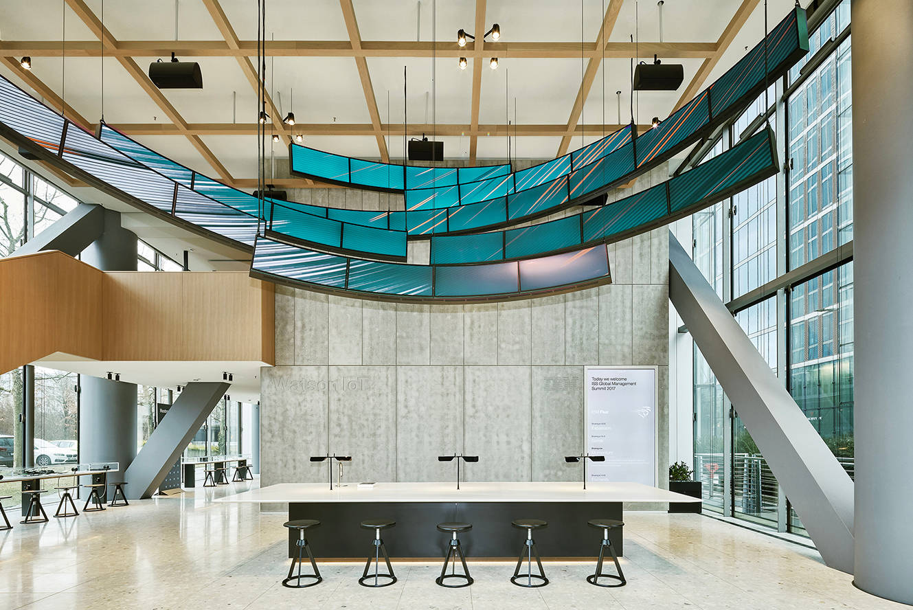 Modern, spacious lobby with high ceilings, a grid-like wooden framework, large blue-tinted glass panels, a long central table with stools, and floor-to-ceiling windows letting in natural light.
