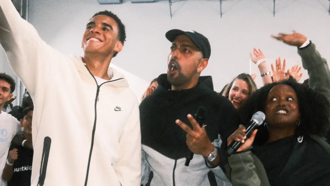 A group of people pose for a cheerful selfie indoors. The person on the left smiles while holding a phone, the middle person flashes a peace sign, and the person on the right holds a microphone. All are surrounded by others celebrating.
