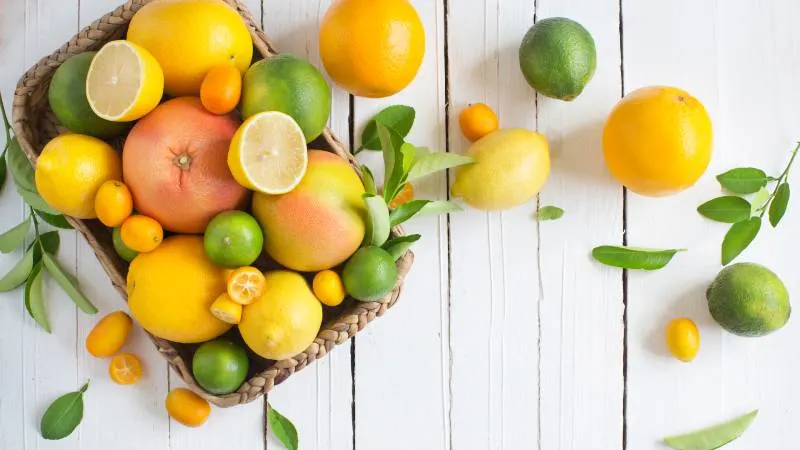 A basket filled with assorted citrus fruits including lemons, limes, grapefruits, and kumquats, sits on a white wooden table. More citrus fruits and green leaves are scattered around the basket.
