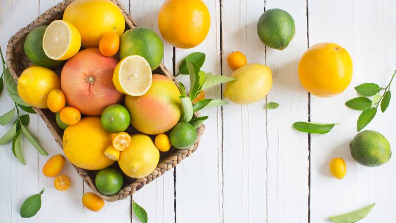 A basket filled with assorted citrus fruits including lemons, limes, grapefruits, and kumquats, sits on a white wooden table. More citrus fruits and green leaves are scattered around the basket.