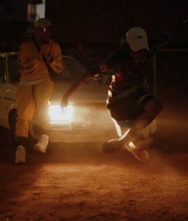 Two people are in front of a car with headlights on at dusk; one person jumps energetically, kicking up dust, while the other, dressed in yellow, claps and watches enthusiastically.