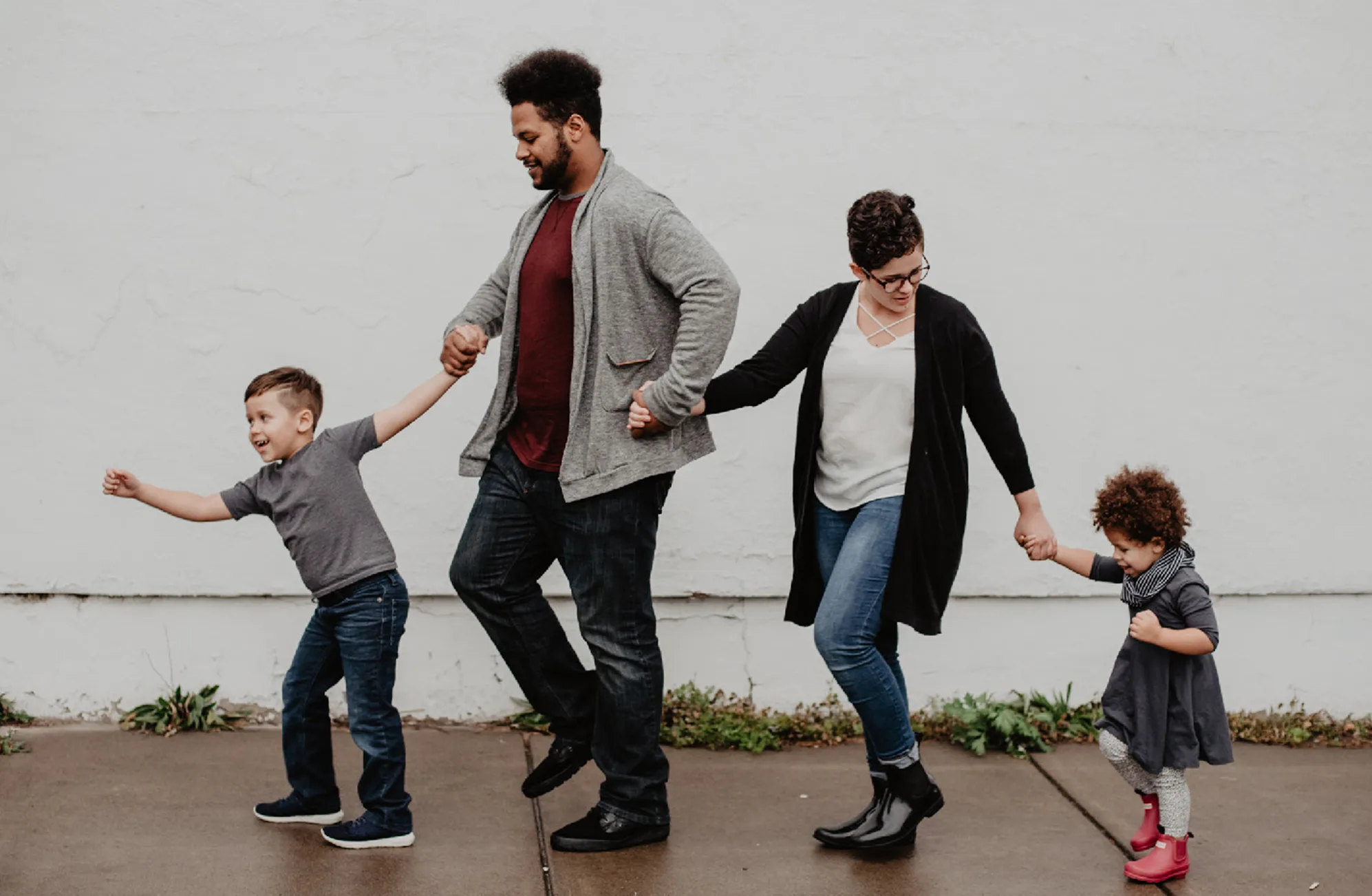 A smiling family of four walks hand in hand on a pavement. The parents are in the center, holding hands with their young son and daughter as they move forward playfully together.