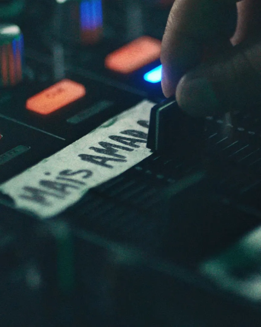 A hand adjusts a slider on a sound mixing console. A piece of tape labeled “Mais Amado” is visible beneath the slider, and various buttons and colored lights surround the control panel.