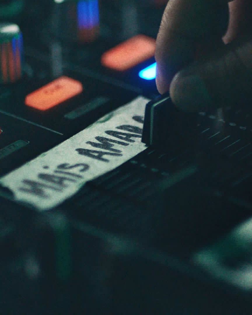 A hand adjusts a slider on a sound mixing console. A piece of tape labeled “Mais Amado” is visible beneath the slider, and various buttons and colored lights surround the control panel.