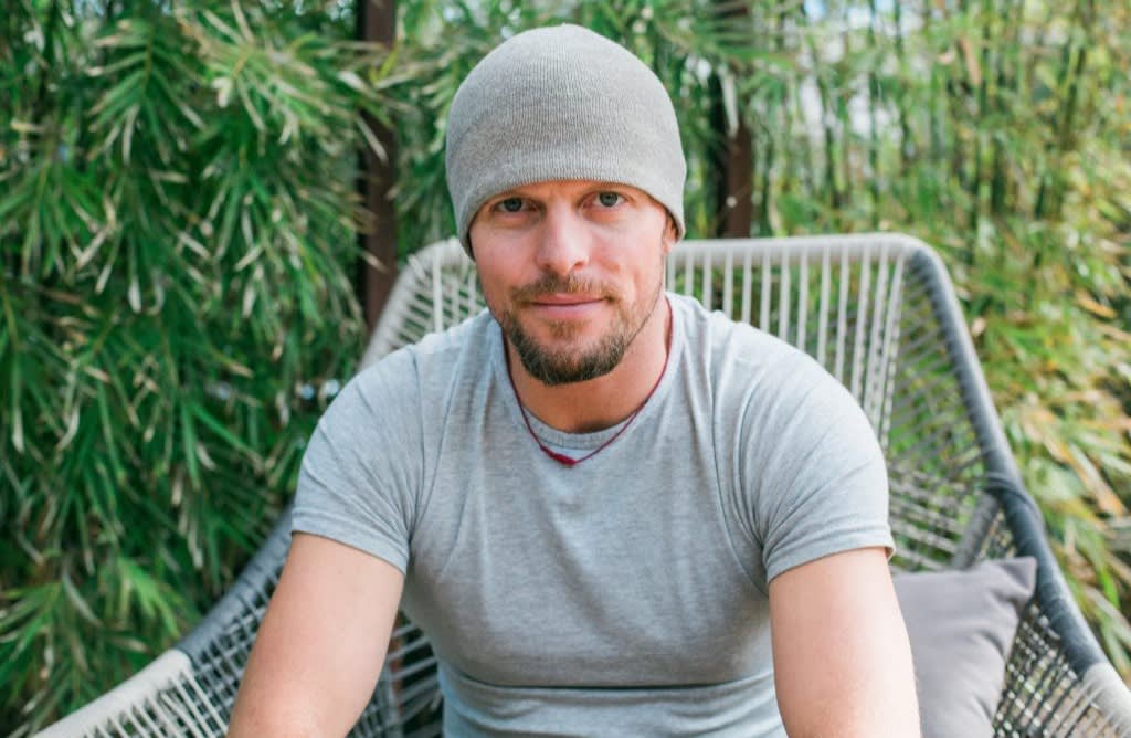 A man with a short beard and mustache wears a gray beanie and a light gray t-shirt while sitting outdoors on a woven chair, surrounded by green foliage.