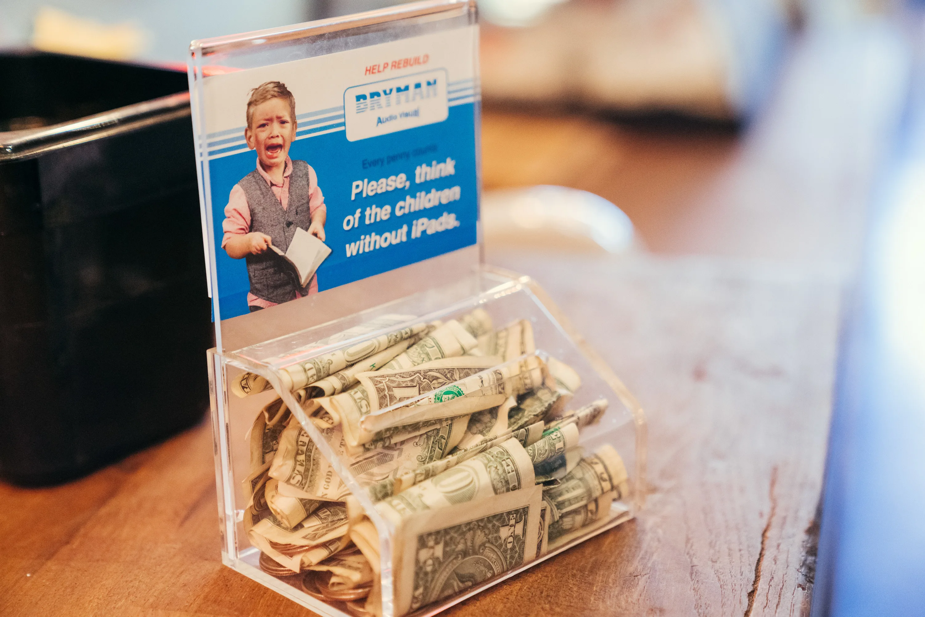 A donation box filled with dollar bills sits on a counter. The sign above it shows a crying child holding paper and reads, Please, think of the children without iPads!.