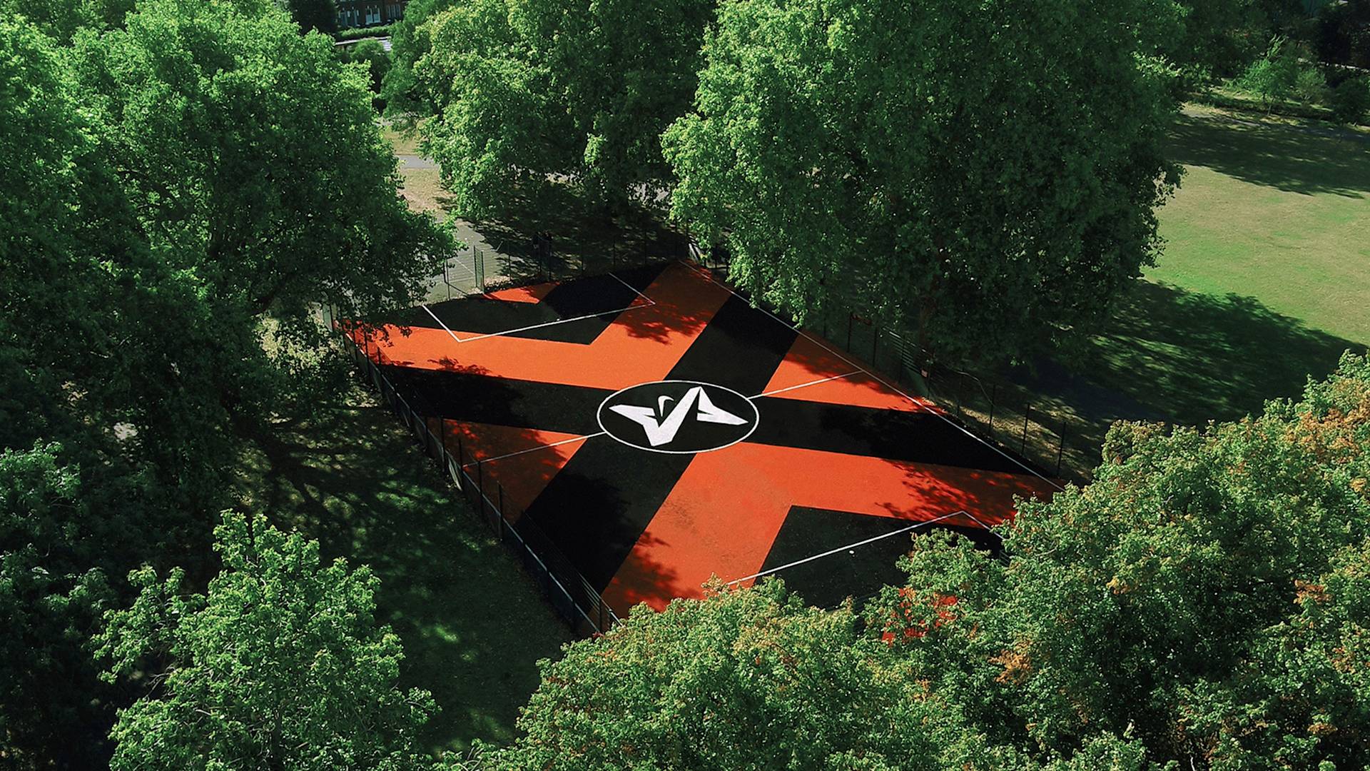 Aerial view of a basketball court surrounded by green trees. The court has a bold black and red geometric pattern with a white logo at the center. Bright sunlight creates contrasting shadows on the court and surrounding grass.