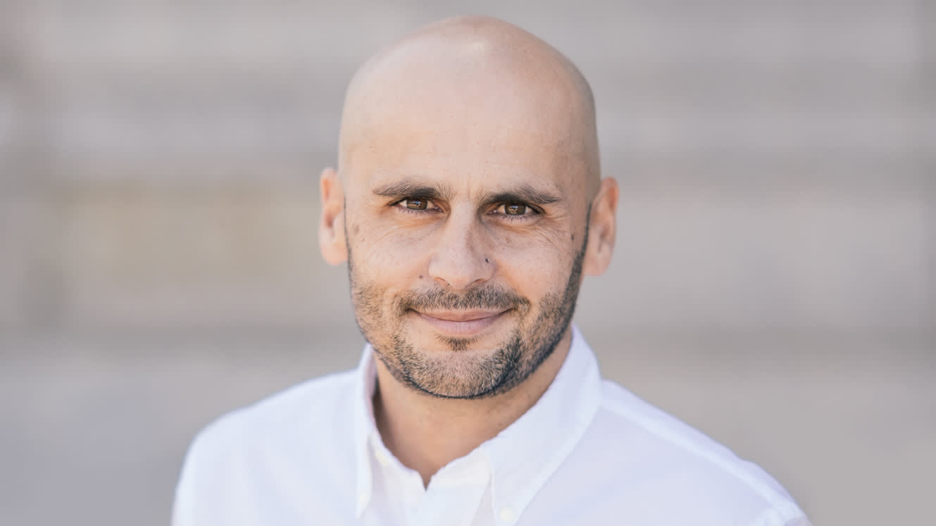 A bald man with a trimmed beard and mustache, wearing a white button-up shirt, smiles softly while looking at the camera. The background is blurred and neutral-toned.