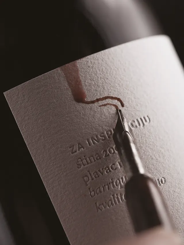 A close-up of a wine bottle label being written on with a fountain pen; red wine drips along the label, partially smudging embossed text.