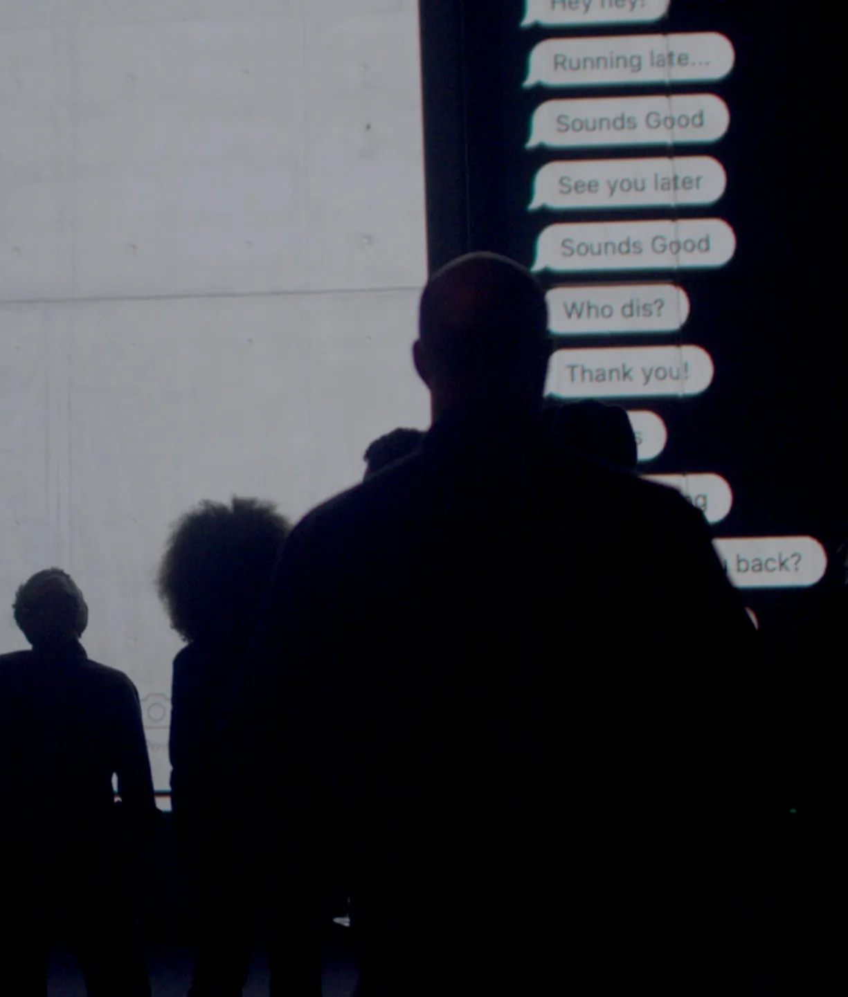 A group of people stand in a dark room, silhouetted against a large screen displaying enlarged text messages in white text bubbles on a black background.