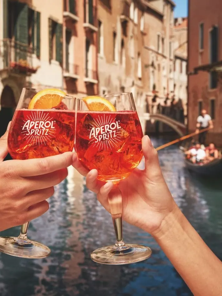 Two hands clink Aperol Spritz glasses with orange slices, in front of a scenic Venice canal with a gondola and colorful buildings in the background.