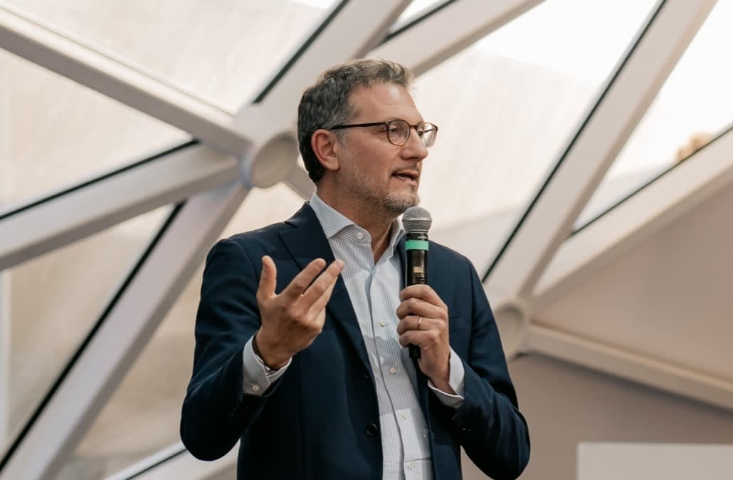 A man in a suit and glasses speaks into a microphone, gesturing with one hand. He stands indoors in front of large windows with geometric metal framing.