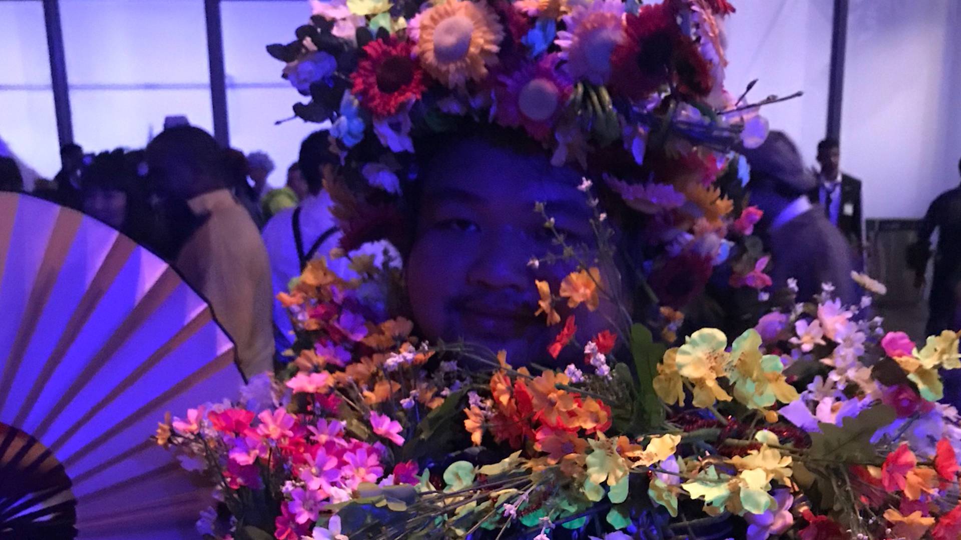 A person wearing a large, colorful floral headdress and garland, surrounded by vibrant flowers, stands in a dimly lit room with a crowd in the background.