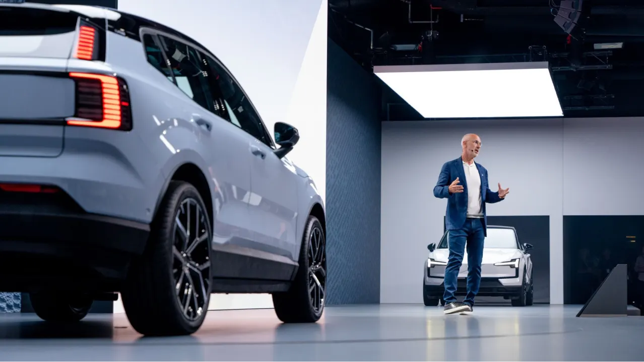 A man in a blue blazer stands on stage presenting, with a white car in the foreground and another white car displayed in the background under bright lighting.