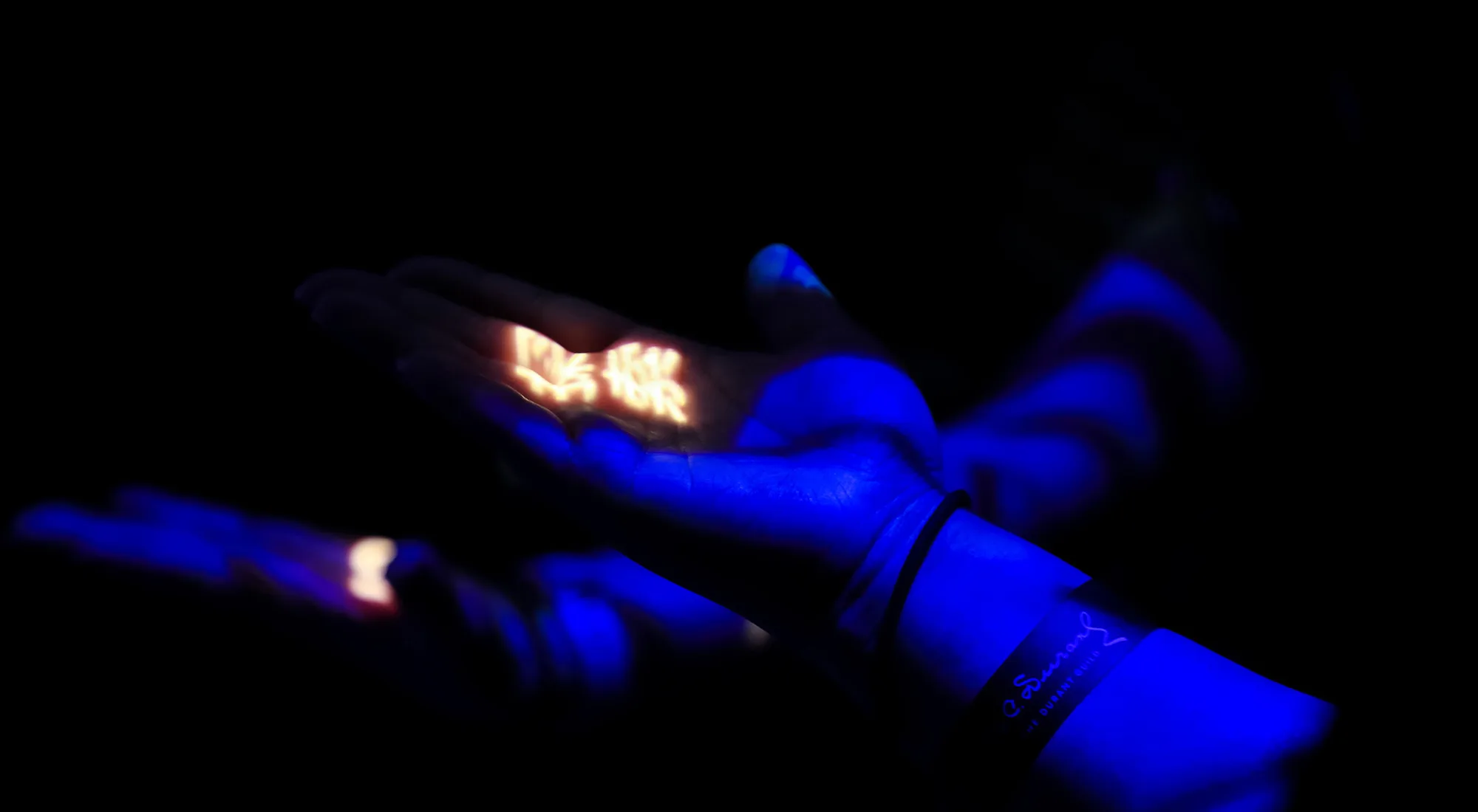 A pair of hands illuminated by blue and purple light, holding a cluster of glowing, warm-colored shapes in the darkness. A bracelet is visible on one wrist.
