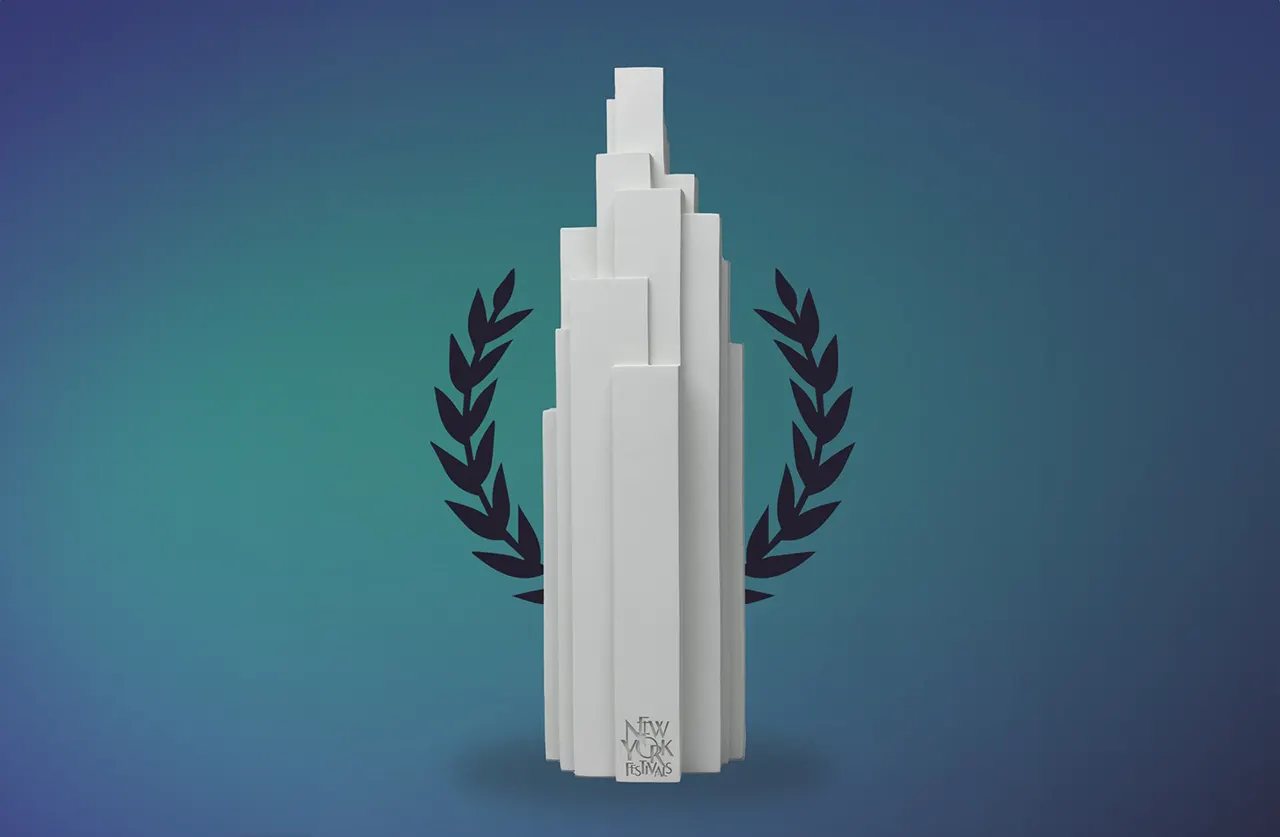 A white, abstract skyscraper-shaped trophy stands upright with black laurel branches behind it, set against a blue gradient background. The base is engraved with “New York Festivals.”.