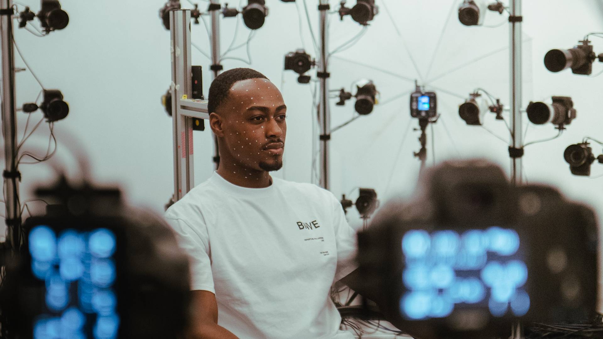A man with tracking dots on his face sits in front of an array of cameras and equipment, possibly for 3D facial scanning or motion capture, wearing a white T-shirt in a studio setting.