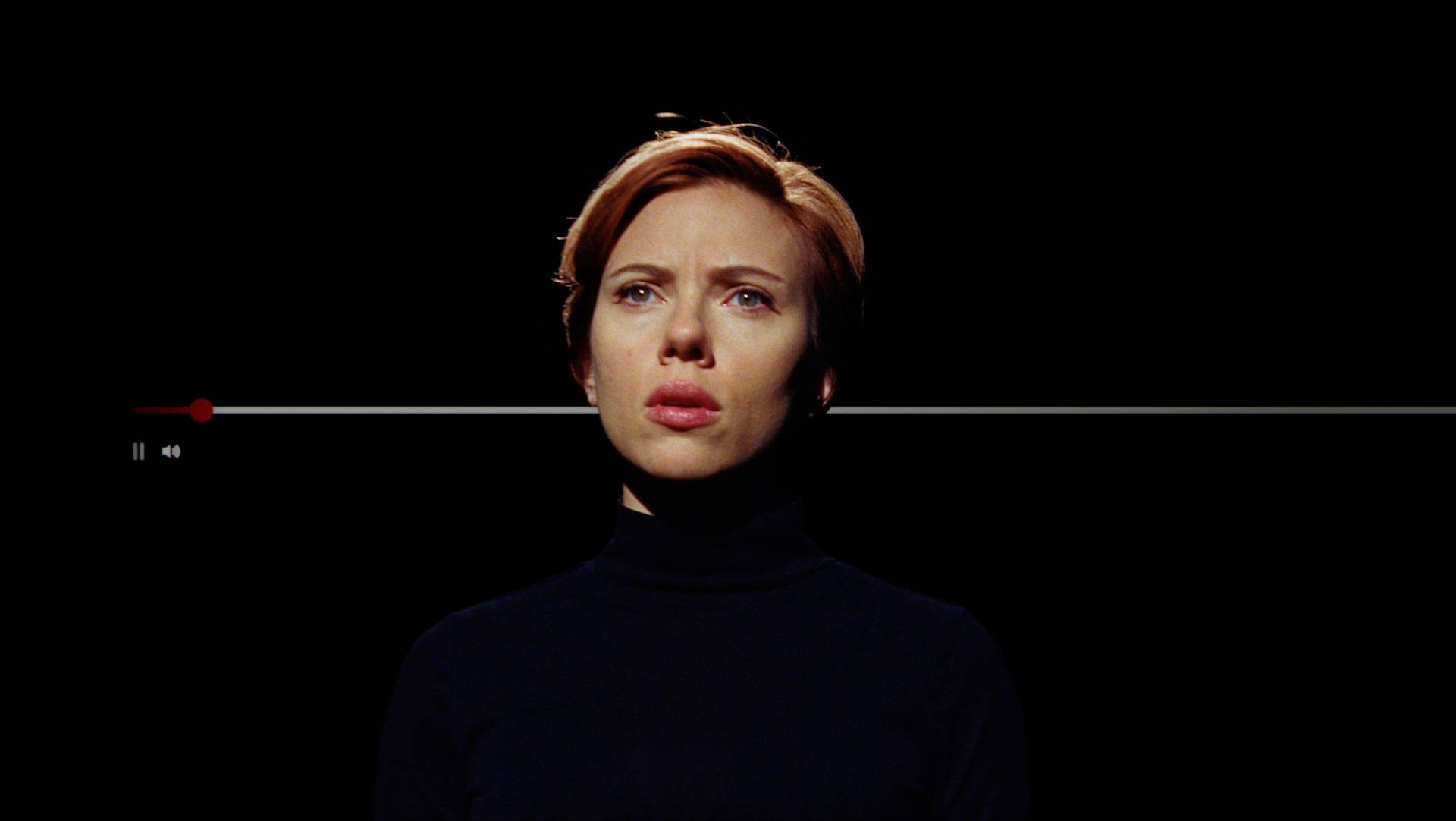 A woman with short red hair and a serious expression is centered against a black background. A video progress bar is superimposed across the image, indicating a paused video.