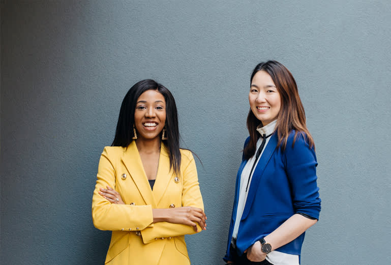 Two women stand smiling in front of a gray wall. One wears a bright yellow blazer with arms crossed; the other wears a blue blazer and white blouse with hands in pockets. Both appear confident and professional.
