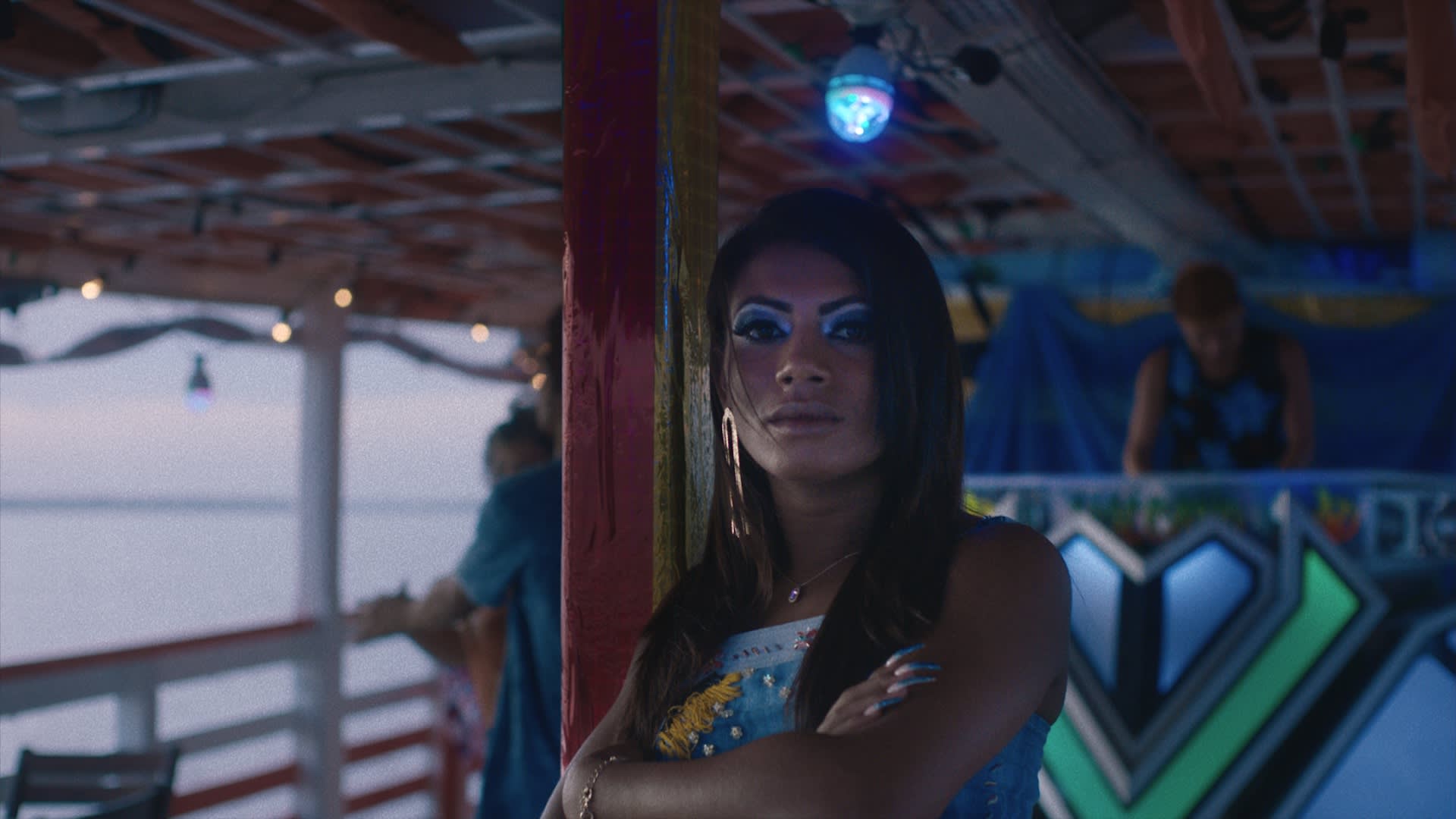 A person with long dark hair, blue eyeshadow, and a confident expression stands with arms crossed on a boat decorated with lights and colorful graffiti, as water and sky are visible in the background.