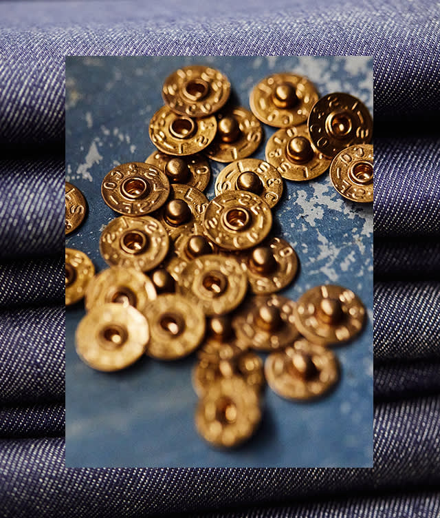 A pile of round brass jean rivets is scattered on a blue textured surface, with folded denim fabric in the background.