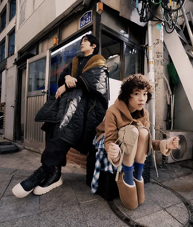 Two people pose on a city sidewalk by a building. One stands, leaning back in a quilted coat; the other crouches, wearing a tan jacket and slippers. Both look away thoughtfully. An address sign and stairway are visible above them.